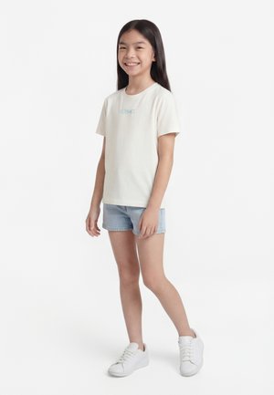 Smilende pige med langt mørkt hår iført en cremefarvet Versace t-shirt, lyseblå denimshorts og hvide sneakers på hvid baggrund.