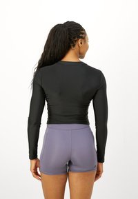 Kvinde med flettet hår i en hestehale iført en sort tætsiddende langærmet top og lilla træningsshorts, vendt væk mod en hvid baggrund.