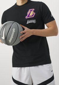 Svart kortärmad t-shirt med en lila och gul Los Angeles Lakers-logotyp och vit text. Bäraren håller en grå basketboll.
