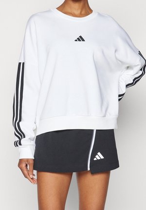 Malli käyttää valkoista pitkähihainen collegepaitaa, jossa on mustat raidat hihoissa, ja mustaa hametta, jossa on valkoiset Adidas-logot ja reunukset.