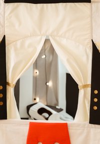 Palapinės žaidimų namelis su balta audinio danga, juodais akcentais ir aukso detalėmis. Yra užuolaidėlės angos ir ryškiai raudonas apvadas apačioje.