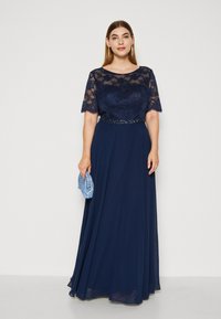 Rochie de seară bleumarin cu mâneci scurte din dantelă, detaliu cu mărgele la talie și fustă vaporoasă, asortată cu o poșetă albastră.