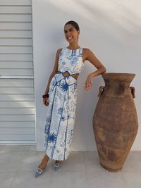 Rochie albă cu modele albastre, cu decupaj la talie și fustă evazată. Pantofi gri cu vârf ascuțit și accente decorative. Ghiveci rustic în apropiere.