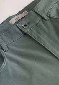 Pantalon vert slim fit avec fermeture à bouton métallique, passants de ceinture et étiquette visible de la marque NEXT à l'intérieur de la ceinture.