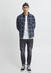 Camisa de franela a cuadros azul y amarillos, camiseta blanca debajo, jeans ajustados negros y zapatillas negras; tejido texturizado y dos bolsillos de parche visibles.