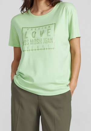 Kobieta w jasnooliwkowej koszulce z napisem "Passion Love MOS MOSH Jeans Heart" w połączeniu z oliwkowymi spodniami, ręce w kieszeniach.