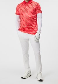 Polo de patrón rojo con cuello, combinado con pantalones blancos y zapatillas blancas. La camisa presenta un diseño geométrico en una tela brillante.
