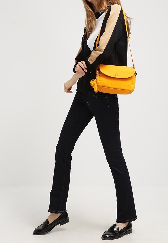 Femme portant une veste noire à manches beiges, un pantalon noir, des mocassins noirs et un sac bandoulière jaune vif.
