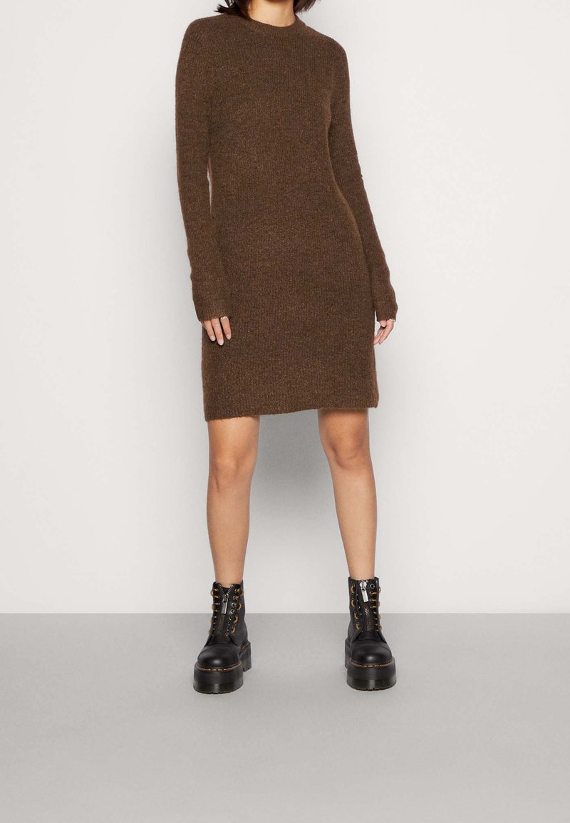 Femme portant une robe en maille marron à manches longues, associée à des bottes à plateforme noires à lacets, debout devant un fond clair uni.