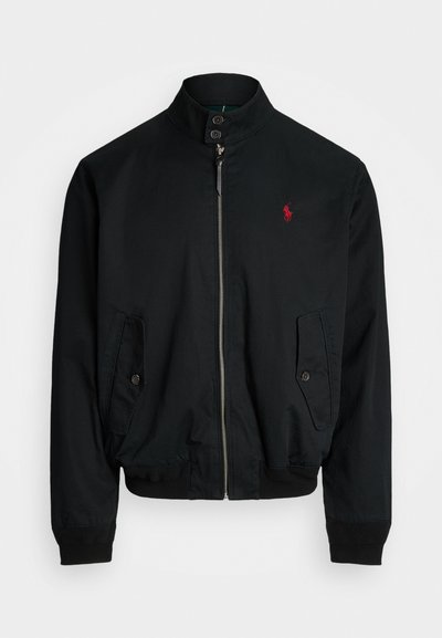 Casaco bombazina preto com gola alta, fecho de correr à frente, dois bolsos laterais, punhos em ribana e um pequeno logotipo vermelho no peito. Textura de tecido suave.