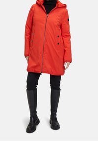 Manteau rembourré rouge avec une capuche, présentant une fermeture éclair à l'avant, des boutons-pression et deux poches latérales. Porté par-dessus des leggings noirs et des bottes noires montantes.