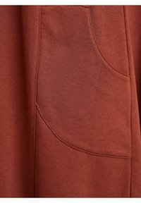 Zbliżenie na materiał w kolorze rdzy z widocznym przeszyciem i zakrzywionym detalem kieszeni na ubraniu.