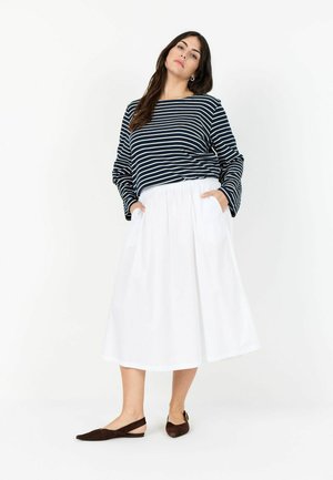 Kobieta w granatowo-białej pasiastym topie z długim rękawem, białej spódnicy do kolan z kieszeniami oraz ciemnobrązowych balerinach na pasku, stojąca na tle białego tła.