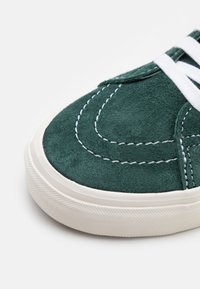 Dedão de sneaker em camurça verde com costura branca. Apresenta uma sola de borracha texturizada e atacadores brancos. Superfície lisa e fosca com costuras detalhadas.