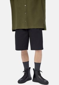 Jambes portant un short noir arrivant au genou, des chaussettes gris foncé, des bottes noires à lacets et une chemise verte à boutons dépassant la taille.