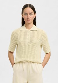 Polo en tricot couleur crème clair avec des manches courtes, un motif texturé, un col classique et une patte de boutonnage à cinq boutons sur le devant. Tissu doux.