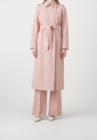 Vaaleanpunainen trenssitakki kaksirivisillä nappeilla, vyötäröllä solmittava vyö ja pitkät hihat, yhdistettynä vastaaviin leveälahkeisiin housuihin ja teräväkärkisiin kenkiin.