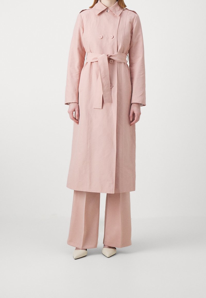 Vaaleanpunainen trenssitakki kaksirivisillä nappeilla, vyötäröllä solmittava vyö ja pitkät hihat, yhdistettynä vastaaviin leveälahkeisiin housuihin ja teräväkärkisiin kenkiin.