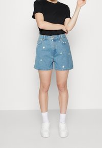 Hellblaue Jeansshorts mit weißer Gänseblümchen-Stickerei, hoher Taille und weitem Schnitt. Kombiniert mit einem schwarzen T-Shirt und weißen Turnschuhen.