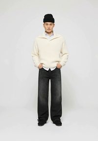 Jersey de punto acanalado de color crema con un collar ancho, superpuesto a una camisa de rayas azul y blanca. Jeans oscuros de corte holgado y zapatos negros.