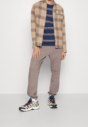 Chemise à carreaux beige et noire sur un t-shirt rayé bleu marine. Pantalons de survêtement marron clair et baskets blanches avec des accents violets et dorés.
