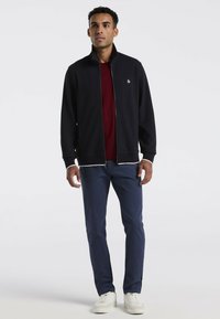 Marineblauwe ritsjacket met hoge kraag, gecombineerd met een bordeauxrood shirt, blauwe slim-fit broek en witte sneakers. Gladde stof met subtiele textuur.