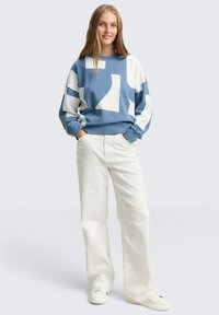 Jonge vrouw staat met handen in haar zakken, gekleed in een sweatshirt met blauw-witte abstracte patronen en witte wijde broek met witte sneakers.