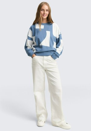 Jonge vrouw staat met handen in haar zakken, gekleed in een sweatshirt met blauw-witte abstracte patronen en witte wijde broek met witte sneakers.