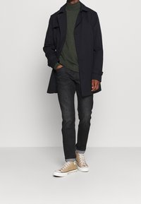 Dlouhý námořnicky modrý trenčkot přes tmavě zelený rolák, doplněný tmavými džínami a béžovými vysokými teniskami s bílými podrážkami a tkaničkami.