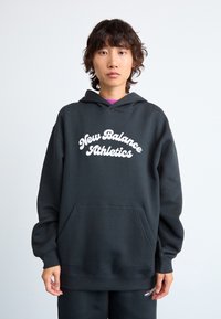 Personne avec des cheveux courts et ondulés portant un hoodie noir New Balance Athletics et un pantalon assorti devant un fond clair.