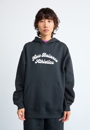 Personne avec des cheveux courts et ondulés portant un hoodie noir New Balance Athletics et un pantalon assorti devant un fond clair.