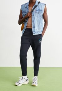 Jeansväst utan ärmar, brun linne, svarta joggers med turkosa Nike-loggan, vita sneakers med svarta detaljer, står på grön golv.