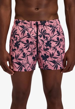 Parte superiore del corpo e gambe maschili che indossano pantaloncini da bagno rosa con motivo nero di foglie e fiori tropicali e coulisse nera in vita.