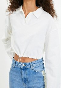Polo bianco a maniche lunghe, corto, con colletto bottonato ed orlo elasticizzato, abbinato a jeans in denim azzurri a vita alta.