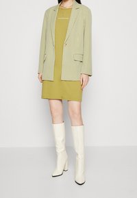 Grün-weiß kariertes Sakko über einem khakifarbenen Kleid, ergänzt durch kniehohe weiße Stiefel mit quadratischer Spitze und Blockabsätzen.