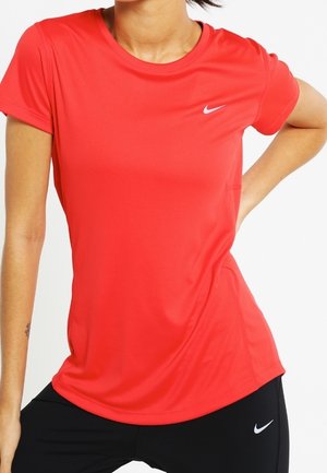 Vrouw draagt een rood Nike T-shirt met korte mouwen en zwarte Nike broek, staand met één hand op haar heup tegen een witte achtergrond.