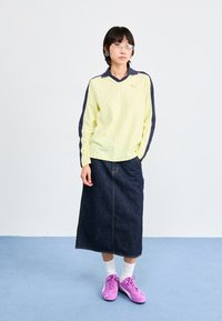 Chemise à manches longues rayée jaune avec des accents navy, associée à une jupe en denim foncé et des baskets roses. Le mannequin se tient devant un fond bleu clair.