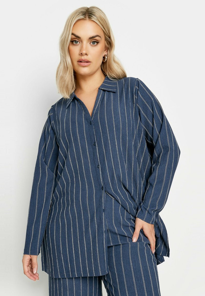 Camicia a maniche lunghe blu navy a righe, con colletto e vestibilità ampia. Presenta righe bianche verticali e un dettaglio con spacco laterale.