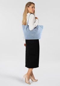 Borsa a tote in suede azzurro chiaro con texture morbida, dotata di doppio manico e un logo discreto. Indossata da un modello con una maglietta bianca e una gonna nera.