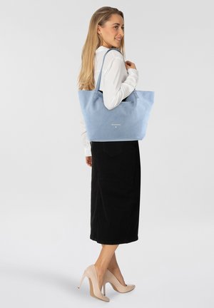 Sac fourre-tout en daim bleu clair avec une texture douce, doté de bretelles doubles et d'un logo discret. Porté par un mannequin avec un haut blanc et une jupe noire.