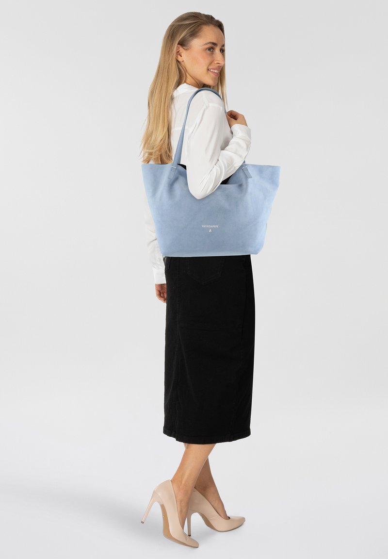 Borsa a tote in suede azzurro chiaro con texture morbida, dotata di doppio manico e un logo discreto. Indossata da un modello con una maglietta bianca e una gonna nera.