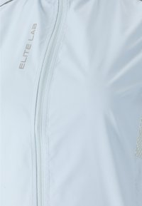 Lyseblå atletisk jakke med fuld lynlås, åndbare mesh-detaljer og "ELITE LAB" logo i sølv på forsiden.