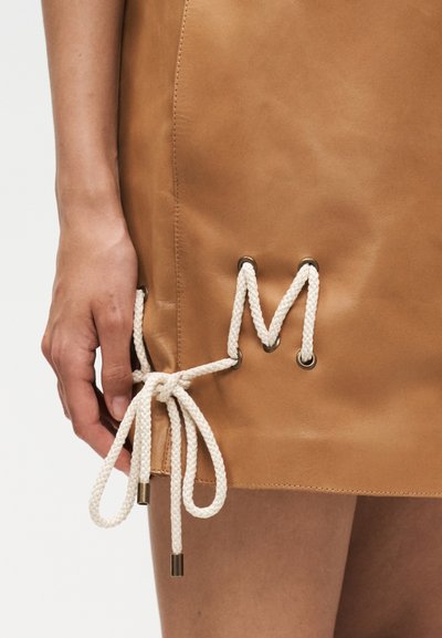 Χέρι δένει λευκά πλεκτά κορδόνια μέσα από μεταλλικά κουμπιά στο πλάι μιας ταμπά δερμάτινης φούστας.