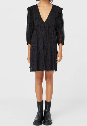 Kobieta w czarnej sukience do kolan z przezroczystego materiału w groszki, z głębokim dekoltem w serek, bufkami na rękawach 3/4 oraz czarnych botkach za kostkę.