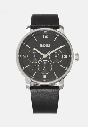 Reloj con correa de cuero negro, caja redonda de plata, esfera negra con tres subesferas y marcadores y manecillas de horas en plata.