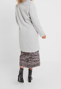 Gray coat with a structured design, long sleeves, and a tailored fit. Worn over a patterned, knee-length skirt and black ankle boots.