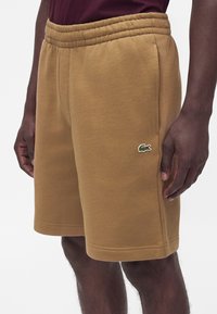 Homme portant un short beige jusqu'aux genoux avec une taille élastique et un petit logo crocodile vert sur la zone de la poche latérale.