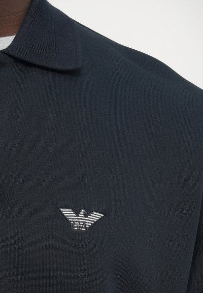 Námornícka polokošeľa s plochým golierom, na ľavej strane hrude s vyšívaným emblémom orla s pruhmi. Materiál je hladký a textúrovaný.