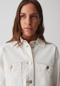 Giacca in denim bianco con collo a collare, bottoni in metallo argentato e due tasche frontali. Texture liscia con dettagli di cucitura visibili.