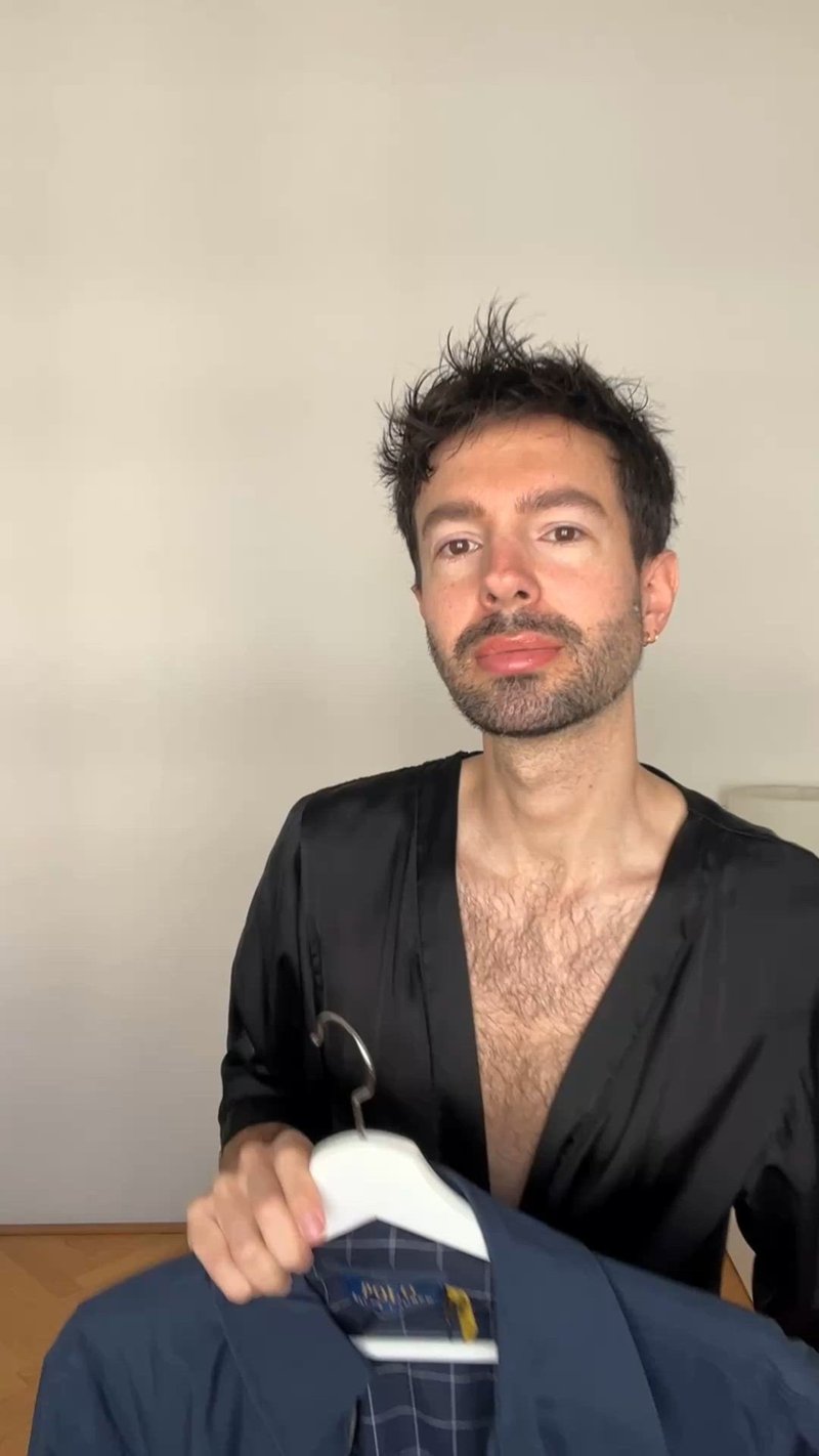 Robe en satin noir avec un décolleté en V profond, maintenue ouverte, révélant une chemise à carreaux bleus sur un cintre blanc. Le modèle a les cheveux courts et une barbe.
